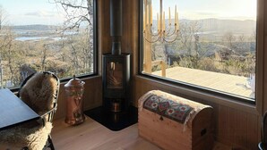 Ein gemütliches Zimmer mit Kaminofen, gepolstertem Sitzplatz, Tisch und großen Fenstern mit schöner Aussicht.