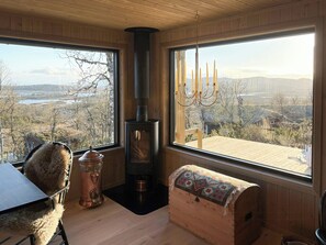 Una habitación acogedora con una estufa de leña, un banco acolchado, una mesa y ventanas amplias que ofrecen una vista panorámica.