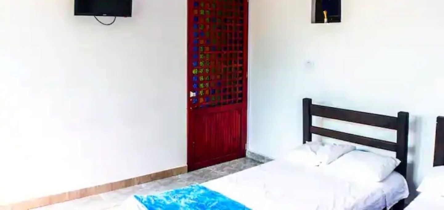 Quarto com uma cama, uma porta vermelha e uma pequena TV fixada na parede.