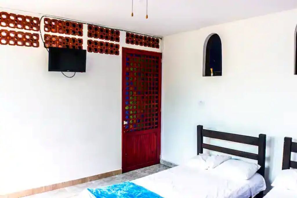 A room with a bed, a TV mounted on the wall, a red door with a colorful grid pattern, and a small window.