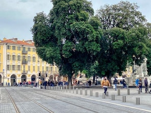 Une place publique avec des voies de tramway, un grand arbre et une statue.
