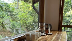 Ein Holztisch mit Kaffeemaschine, Tassen und Teekanne vor einem Fenster mit Blick ins Grüne.