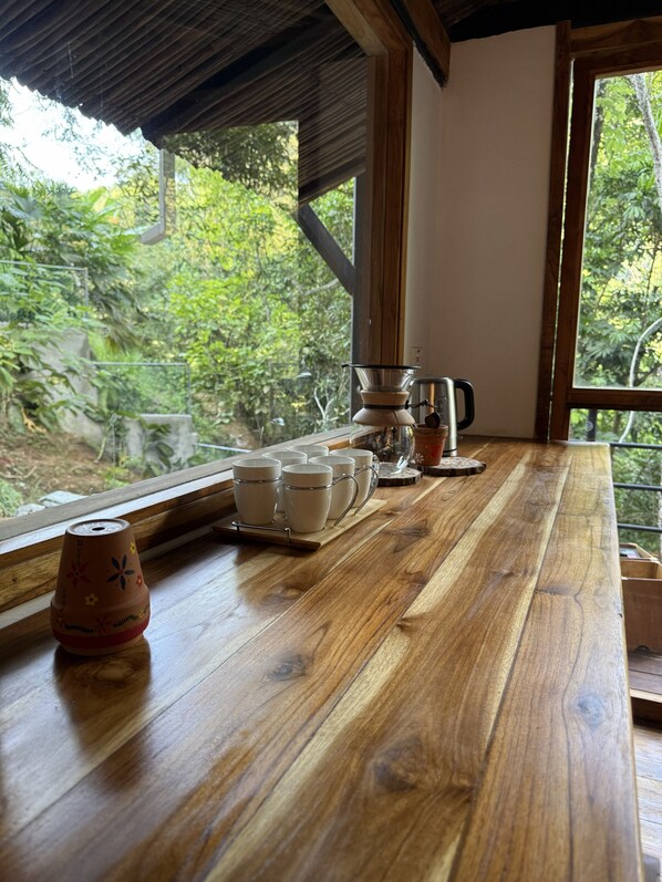 Una mesa de madera con una cafetera, tazas y un hervidor de té junto a una ventana con vistas a la vegetación.