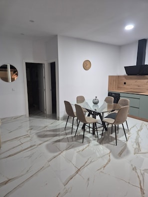 A dining area with a glass table, six chairs, a clock on the wall, and a kitchen with a stove and cabinets. - Lola's Little House (Conil de la Frontera)