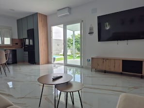 Una sala de estar moderna con televisor de pantalla plana, comedor con sillas, cocina equipada con lavavajillas y puerta corrediza de vidrio que da a un patio.