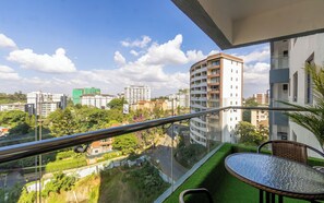 Ein Balkon mit Glasgeländer, Tisch und Stühlen mit Blick auf eine Stadtlandschaft mit Gebäuden und Grünflächen.