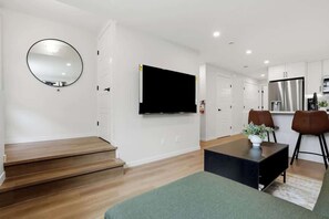 A modern living room with a flat-screen TV, a round mirror, a staircase, a dining area with two chairs, and a kitchen in the background. - New Modern 1Br Condo Near Airport (Calgary)