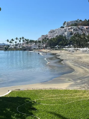 Ein Strand mit blauem Meer, einer grünen Fläche und einer Reihe weißer Gebäude im Hintergrund.