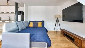 A modern bedroom with a bed, a sofa, a TV, and a kitchen area. - Bastille · 12th · Apartment with Mezzanine – managed by Mey Agency (Paris)
