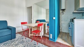 A compact living space with a blue sofa, a dining table with red chairs, a kitchen area with a white refrigerator, and a patterned rug. - Convention · 15th · Colorful & Bright Studio – managed by Mey Agency (Paris)