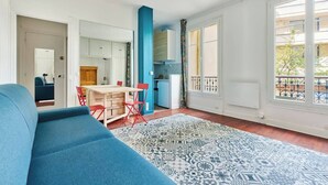 A modern living room with a blue sofa, a dining area with a table and chairs, a kitchen with blue cabinets, and large windows with a view of the outside. - Convention · 15th · Colorful & Bright Studio – managed by Mey Agency (Paris)