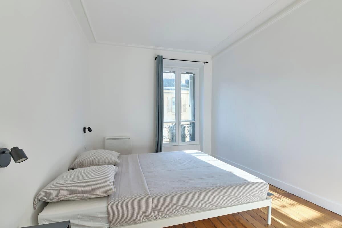 Une chambre à coucher avec un lit, une fenêtre avec des rideaux, une lampe fixée au mur et un radiateur.