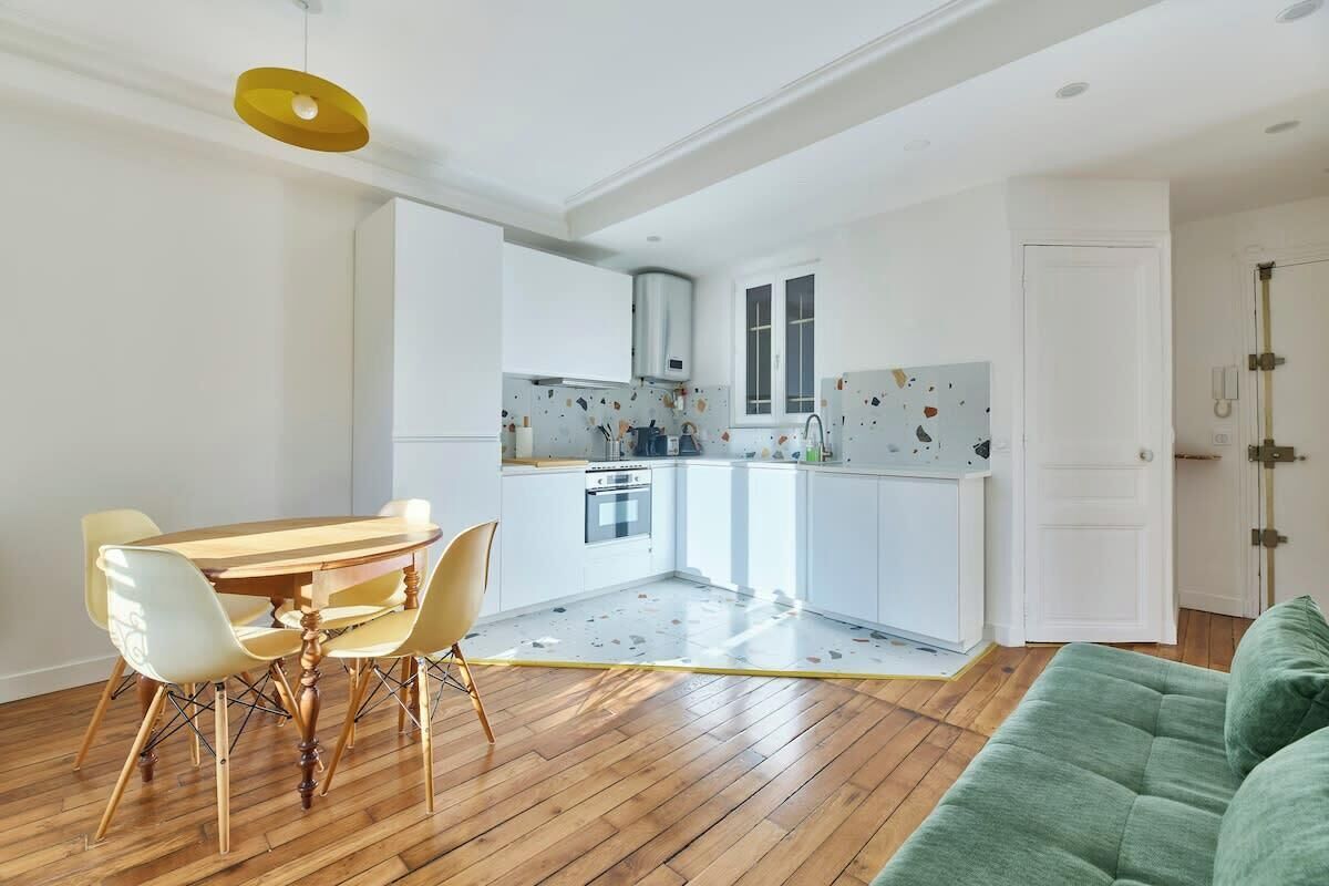 Une cuisine compacte avec des meubles blancs, une table à manger avec des chaises et un canapé vert dans l’espace salon.
