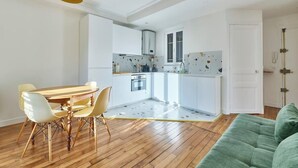 Cocina compacta con armarios blancos, una mesa de comedor con sillas y un sofá verde en la sala.