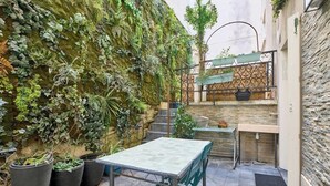 Un pequeño patio al aire libre con una mesa y sillas, rodeado de macetas con plantas y un muro de piedra.