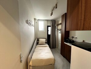 Een smalle hotelkamer met een enkel bed, een kleine kitchenette en een raam met gordijnen.