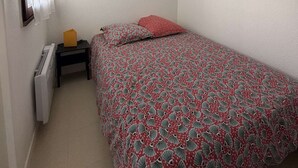 Ein kleines Zimmer mit einem Einzelbett, einer Bettdecke mit Muster, einem Nachttisch mit Lampe und einem Fenster mit Vorhängen.