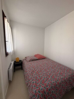 A small, single-bed room with a patterned bedspread, a nightstand with a lamp, and a window with curtains. - Seignosse Océan, one-bedroom apartment near the beach, quiet, pine forest, family of 4 (SEIGNOSSE)