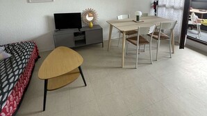 Ein Zimmer mit Bett, Fernseher, Esstisch mit Stühlen, einem kleinen Schrank und einem Fenster mit Vorhängen.