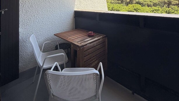 Ein Balkon mit Tisch und zwei Stühlen, einer strukturierten Wand und Blick ins Grüne.