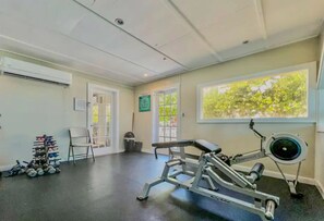 Ein Fitnessraum mit Trainingsgeräten, einem Stuhl, einem Schuhregal und einem Fenster mit Blick auf Bäume.