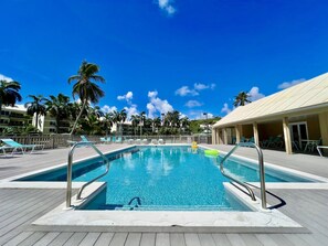 Une piscine avec une terrasse en bois, des palmiers et un bâtiment doté d’un toit de couleur claire.