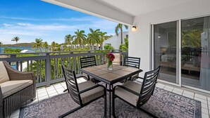 Un balcón con mesa y sillas, un jarrón con flores y vistas a palmeras y una playa.