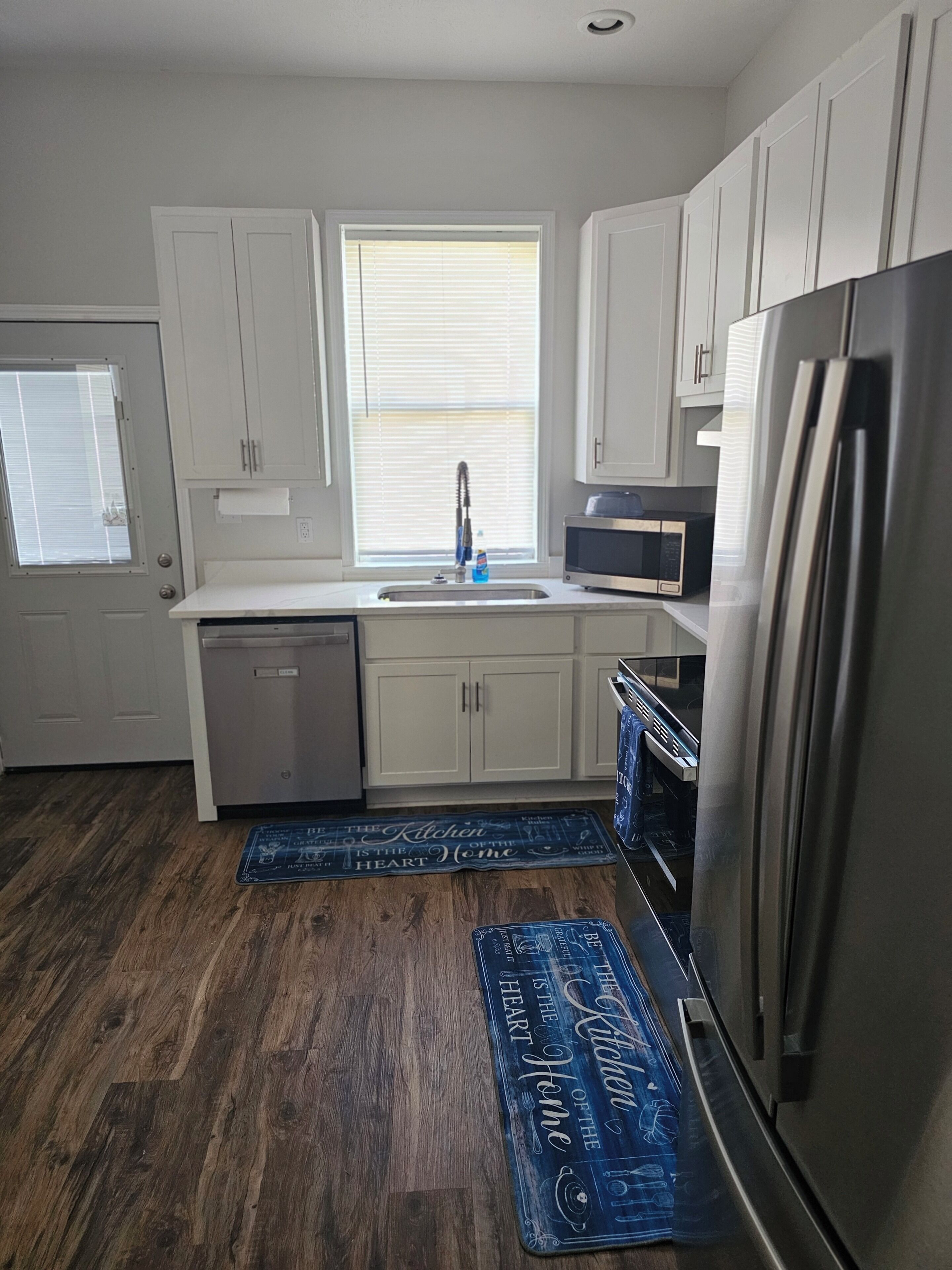 A kitchen with white cabinets, a stainless steel refrigerator, a microwave, and a dishwasher. There is a window with blinds and a wooden floor.