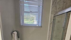 Un baño con inodoro, ducha con puerta de vidrio, ventana con persianas y un cubo de basura.