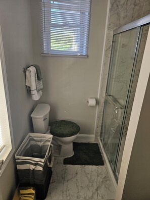 A bathroom with a toilet, a shower with a glass door, a window with blinds, and a trash bin.