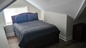 Un dormitorio con techo inclinado, una cama con ropa de cama oscura, una mesita de noche y piso de madera.