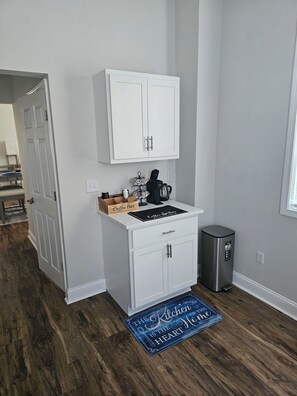 Cocina compacta con armarios blancos, una estación de café y un cubo de basura.
