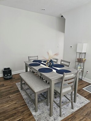 Un comedor con una mesa dispuesta para cuatro personas, bancos, una mesita auxiliar con una cesta y una lámpara fijada a la pared.