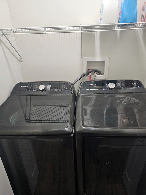 Two black Samsung washing machines side by side with a white wall and a shelf above them.