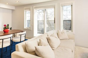 A living room with a white sofa, a wooden dining table, and a blue rug.