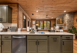 A modern kitchen with wooden cabinetry, a built-in dishwasher, and a stone accent wall. - Cottage at Fern Cove: Couples • Serene • Hot-Tub • Fire-Pit • Close to Downtown (Blue Ridge)