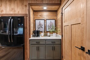 A kitchen with a black refrigerator, two framed botanical prints, a coffee maker, and a potted plant on the counter. - Cottage at Fern Cove: Couples • Serene • Hot-Tub • Fire-Pit • Close to Downtown (Blue Ridge)