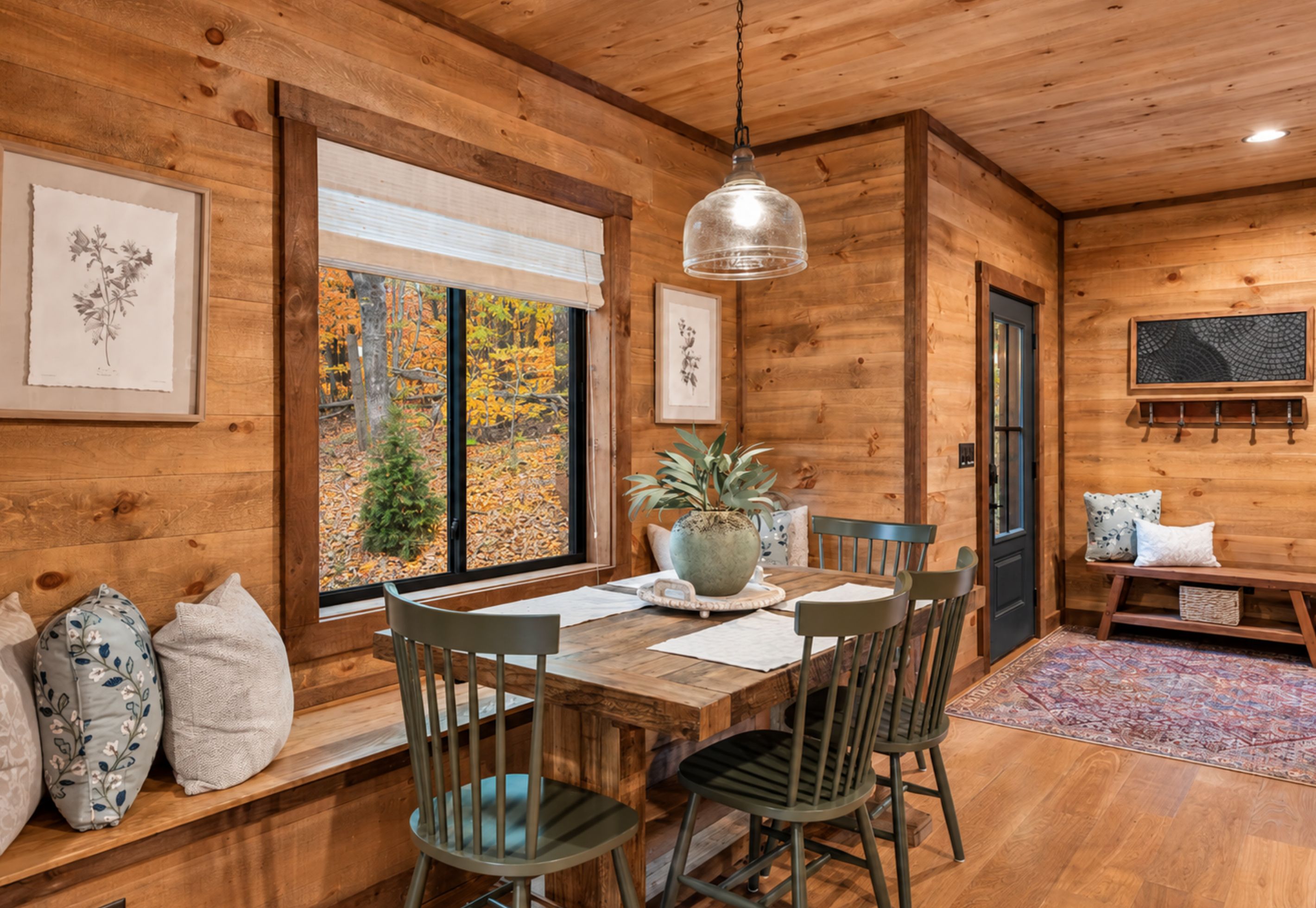 Un intérieur en bois comprenant une table à manger, des chaises, un luminaire suspendu, des œuvres d’art encadrées et une fenêtre donnant sur un paysage automnal.