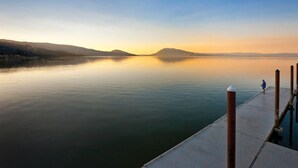 Un lac paisible avec une jetée qui s’avance sur l’eau, une personne debout au bout de la jetée, et des montagnes lointaines sous un ciel dégagé.