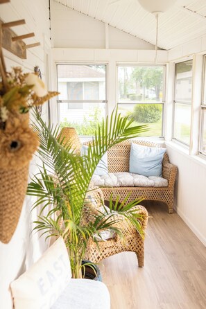 A room with a wicker sofa, a potted plant, and a view through large windows. - The Solo Shore Cottage - Walk to Beach + Sun Porch (Old Lyme)