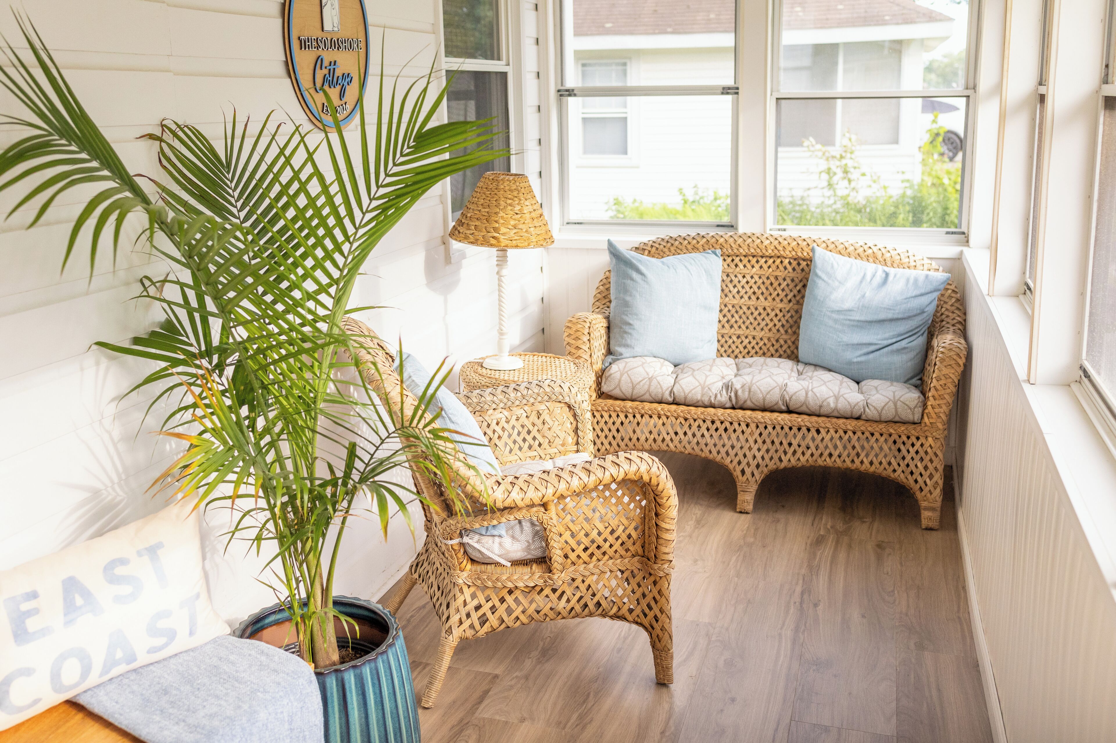 Een rieten bank met kussens, een bijzettafeltje, een lamp en een potplant in een kamer met houten vloeren en een raam.