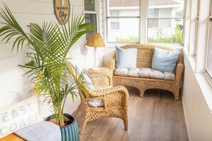 Ein Korbsessel mit Kissen, ein kleiner Tisch, eine Lampe und ein Blumentopf in einem Zimmer mit Holzboden und einem Fenster.
