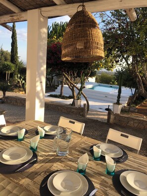Un área de comedor al aire libre con una mesa dispuesta para seis personas, una cesta colgante de mimbre y vistas a una piscina y jardín.