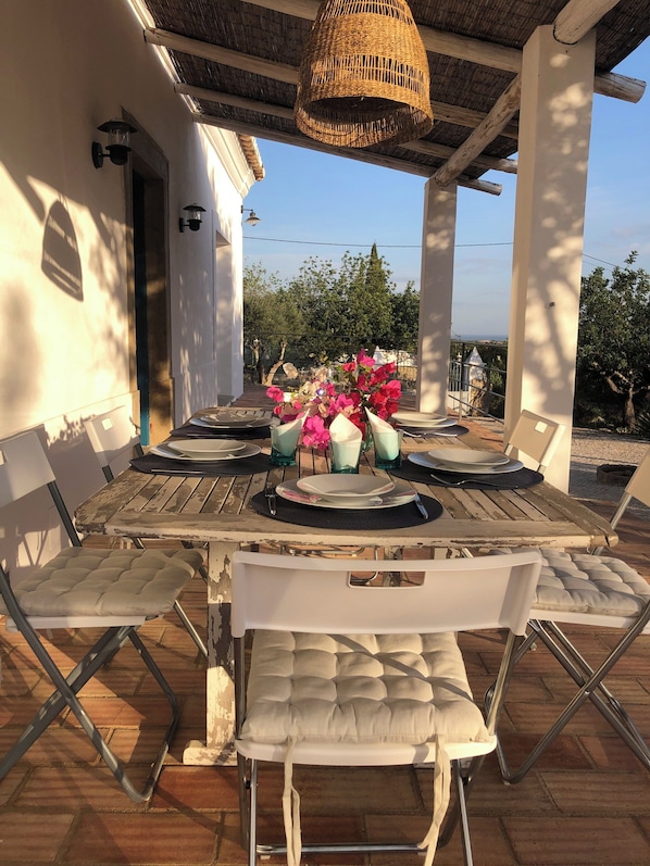A covered outdoor dining area with a table set for two, surrounded by white cushioned chairs.