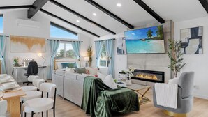 A modern living room with a sofa, dining table, and chairs, a fireplace, and a large TV screen displaying a beach scene.