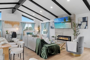 A modern living room with a sofa, dining table, and chairs, a fireplace, and a large TV screen.