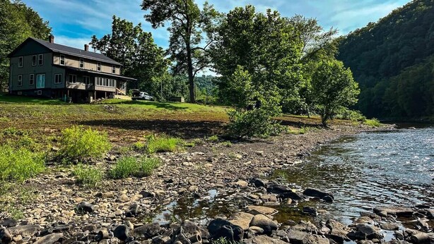 Een pand aan de rivier met een huis, bomen en een rotsachtige oever.