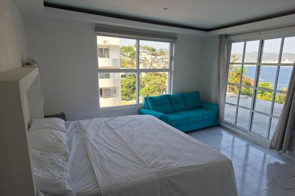 Un dormitorio con cama, sofá color turquesa y amplios ventanales que ofrecen vistas al mar y edificios.