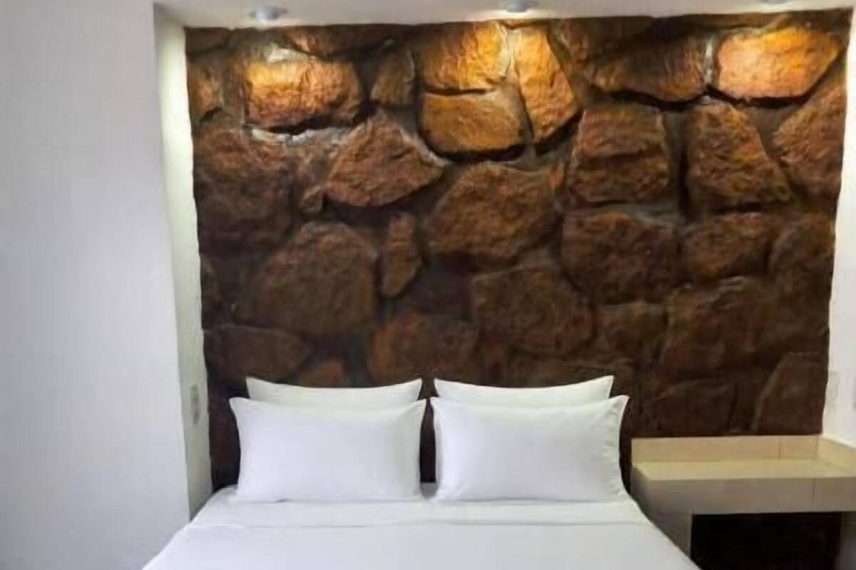 Una cama con ropa blanca, un cabecero de pared de piedra y una mesita de noche con una lámpara.