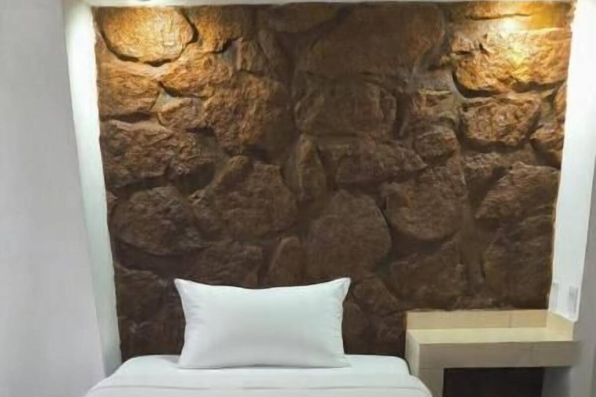 Una cama con colchón y almohadas blancos, un muro de piedra al fondo y un suelo de baldosas de mármol.